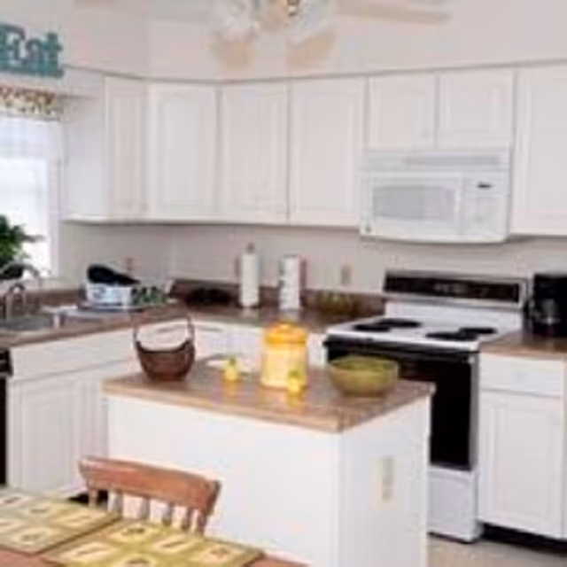A bright kitchen with white cabinets and appliances including a microwave and stove. There is a small island in the center with a basket, a yellow container, and a green bowl on top. A dining table with placemats is partially visible in the foreground.