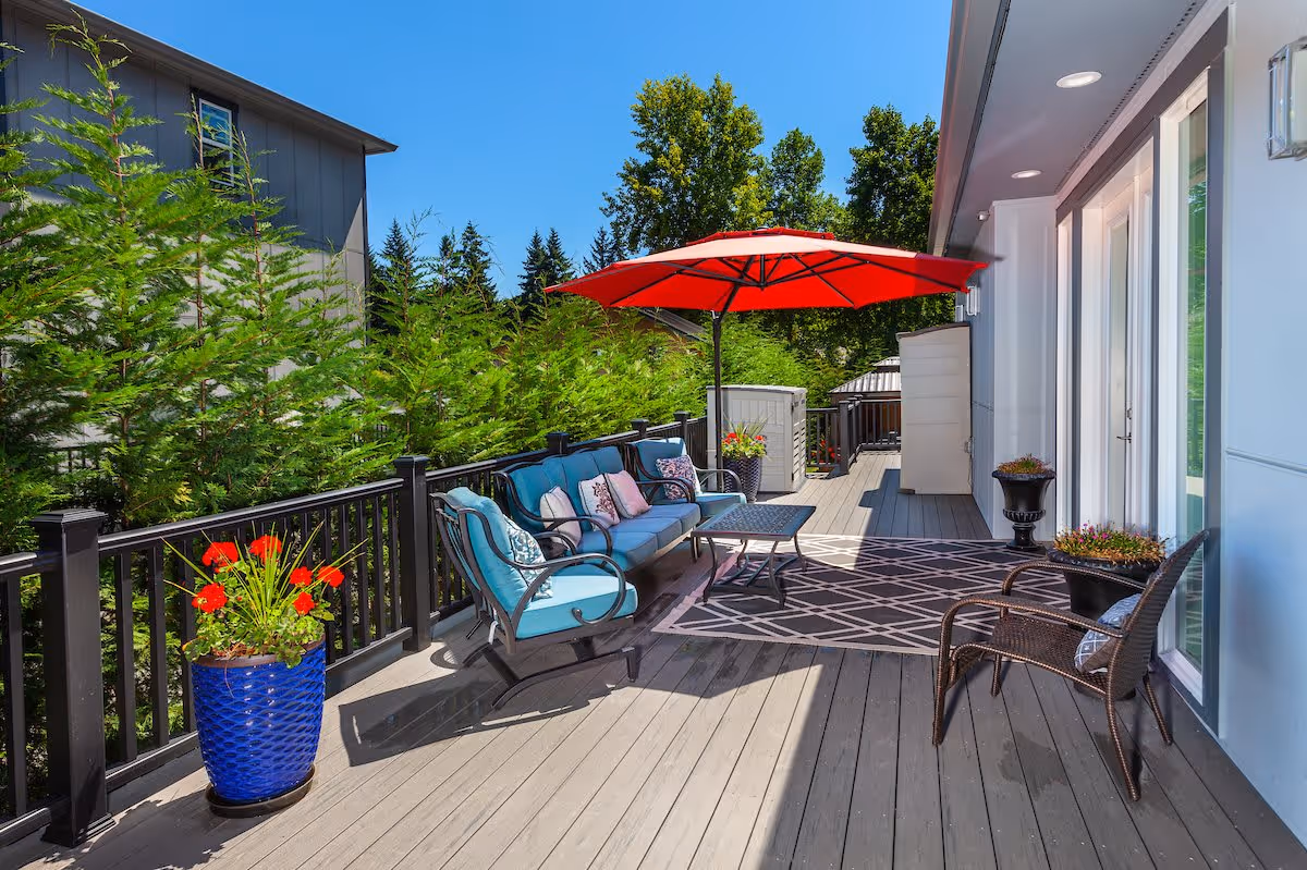Outdoor patio area with a wooden deck featuring a blue cushioned sofa, a matching armchair, a small table, and a large red umbrella providing shade. There are potted plants with red flowers along the railing and near the building. The patio is surrounded by green trees under a clear blue sky.