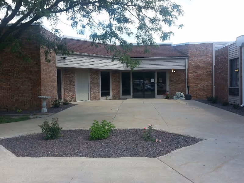 Front exterior view of a single-story brick building with a covered entrance, glass double doors, and a small landscaped area with plants and gravel in front. A tree branch extends over the entrance area.