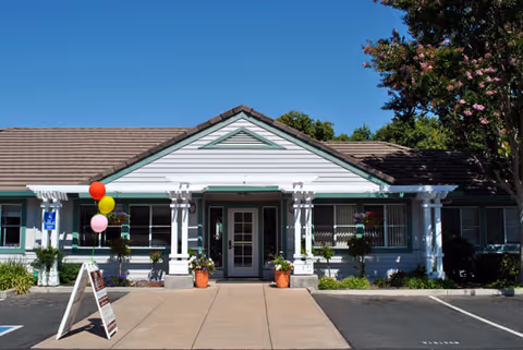 Front exterior view of a single-story building with white siding and green trim, featuring a central entrance with double doors, potted plants on either side, colorful balloons tied to a post, and a sidewalk leading to the entrance under a clear blue sky.