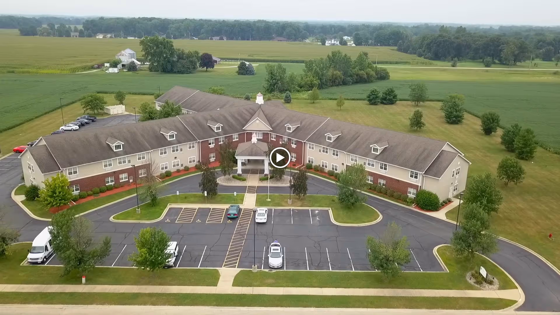 Aerial view of Heritage Woods of Watseka, a large senior living facility building surrounded by parking lots and green fields. The building has a U-shaped layout with a covered entrance and multiple windows. Trees and landscaped areas are visible around the property.