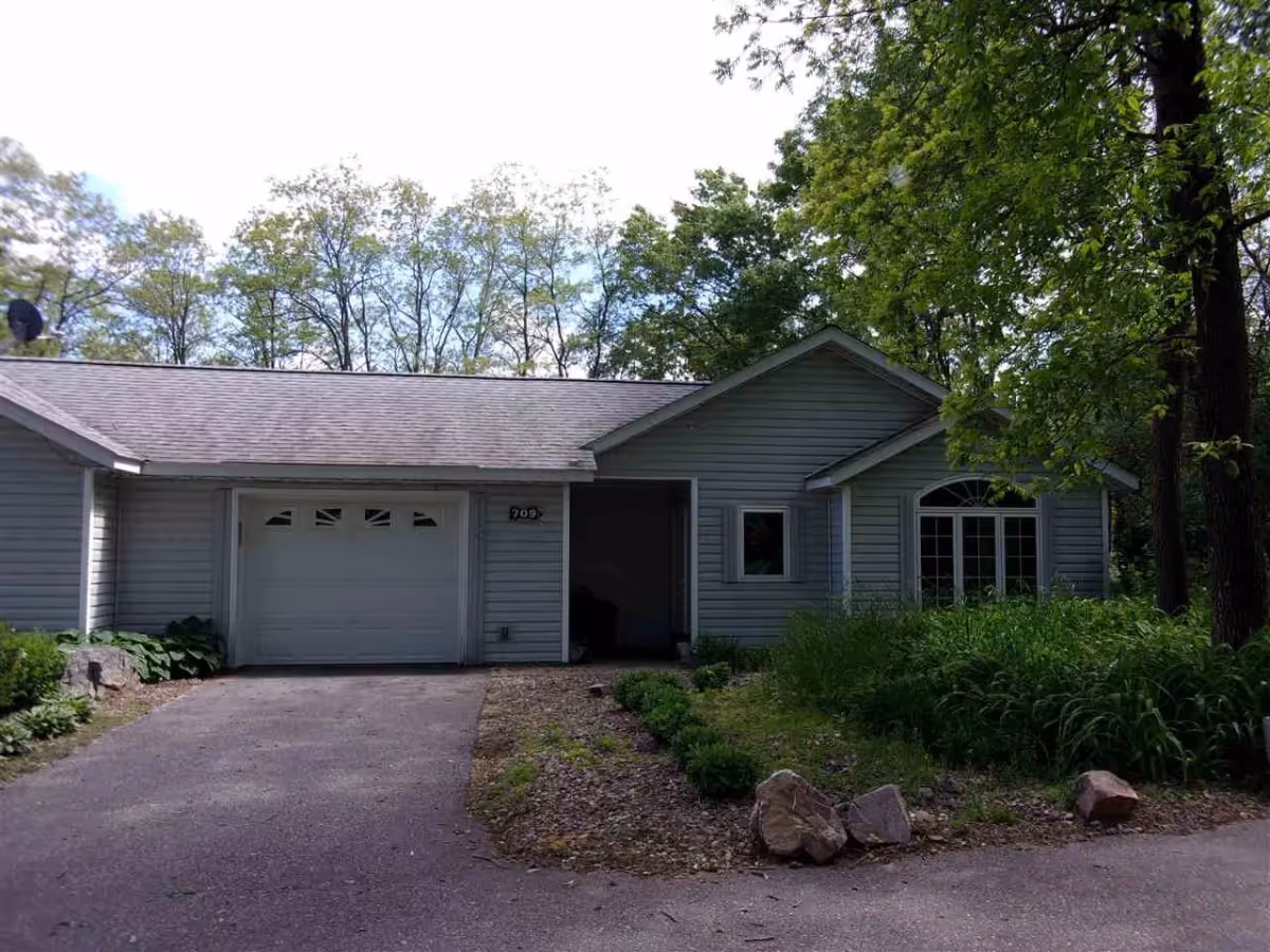 Exterior view of a single-story residential building with gray siding, a garage door, and a driveway. The building is surrounded by trees and greenery, with a garden area featuring rocks and plants in front.
