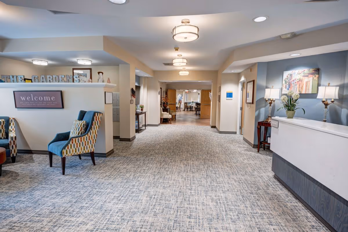 A spacious and well-lit interior hallway of The Gardens at Marysville senior living facility featuring patterned carpet, comfortable armchairs with colorful upholstery, a reception desk with lamps and a flower vase, and a welcoming sign on the wall. The hallway leads to a dining area in the background.