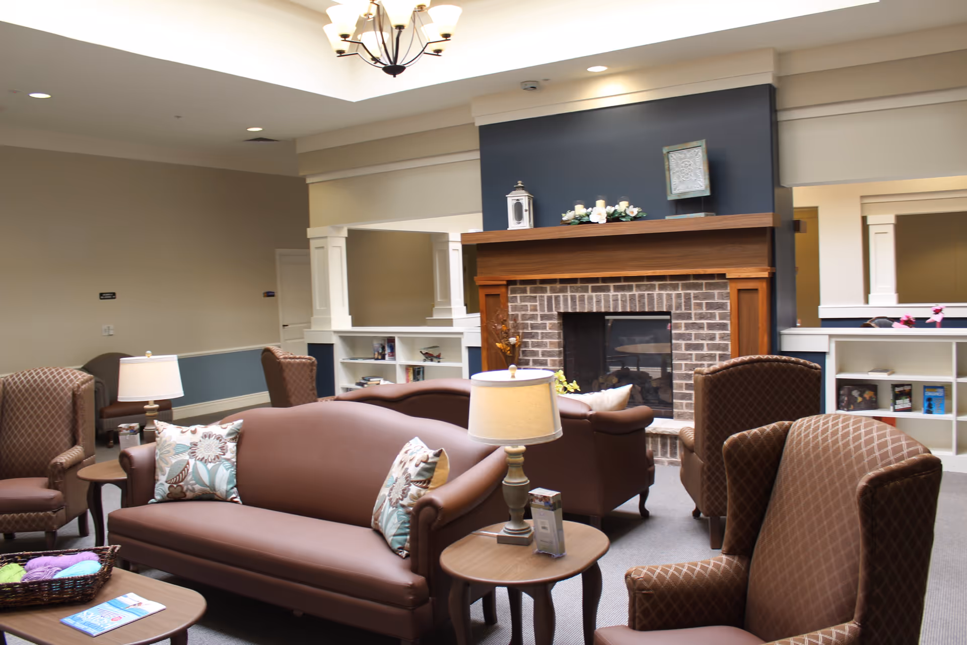 A cozy living room area in a senior living facility featuring a brick fireplace with a wooden mantle decorated with candles and flowers. The room has brown leather sofas and patterned armchairs arranged around small wooden tables with lamps. There are built-in white shelves with books and decorative items, and the walls are painted in neutral tones with a dark accent wall behind the fireplace. The ceiling has recessed lighting and a chandelier.