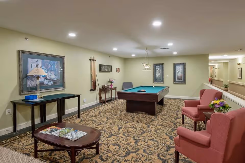 A well-lit recreational room with a pool table in the center, surrounded by patterned carpet. There are two red armchairs with a small table and flower arrangement between them on the right side. On the left side, there is a table with a lamp and a framed picture above it. The walls are painted light green and decorated with framed artwork. Pool cues are mounted on the far wall.