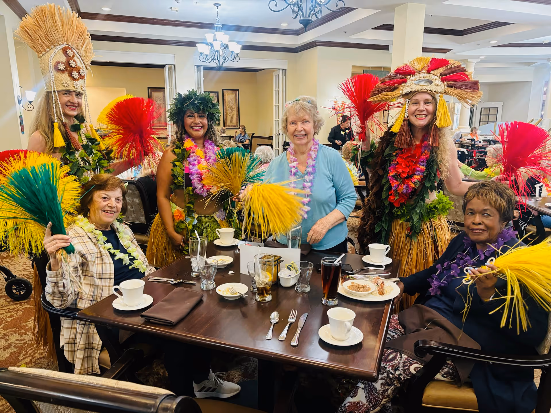A group of elderly women and performers dressed in colorful Hawaiian-themed costumes with grass skirts, leis, and feathered headpieces are gathered around a dining table in a senior living facility. The table has cups, glasses, plates, and utensils, and the setting appears festive and joyful.