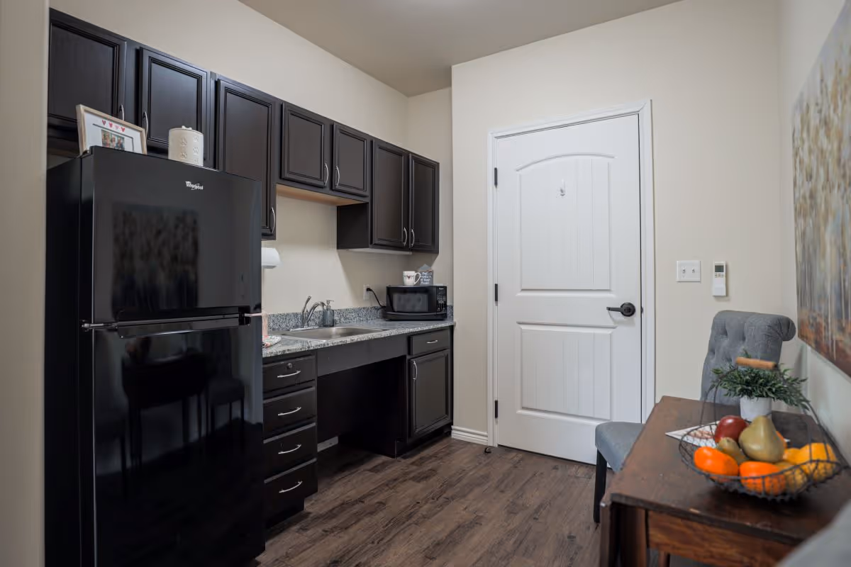 A small kitchen area with dark wooden cabinets, a black refrigerator, a granite countertop with a sink, a microwave, and a wooden table with a gray upholstered chair. On the table is a wire basket filled with various fruits and a small potted plant. A white door is visible in the background.
