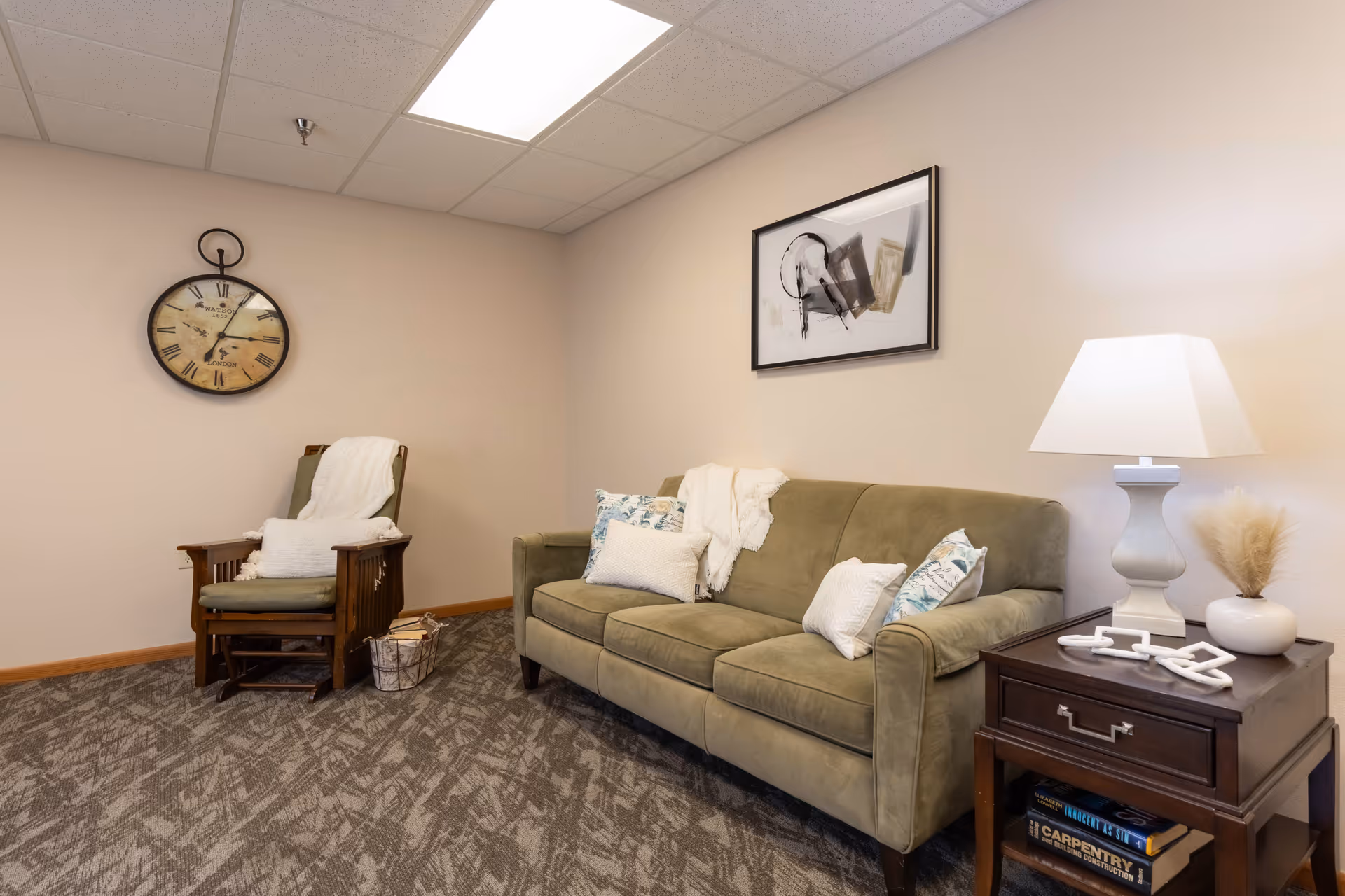 Cozy sitting room with an olive-green sofa, wooden armchair, side table with lamp, wall clock and framed artwork.