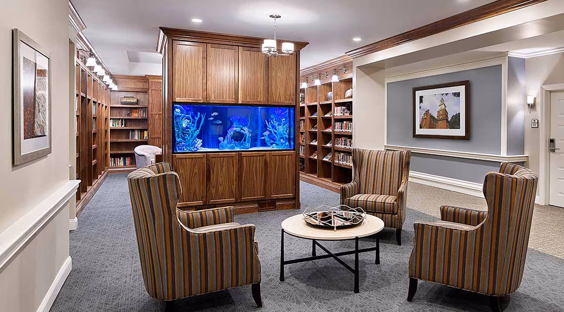 A cozy sitting area in a senior living facility featuring four striped armchairs arranged around a round coffee table with a decorative tray. Behind the seating area is a large built-in wooden bookshelf and a large aquarium with colorful fish. The room has soft lighting, carpeted floors, and framed artwork on the walls.