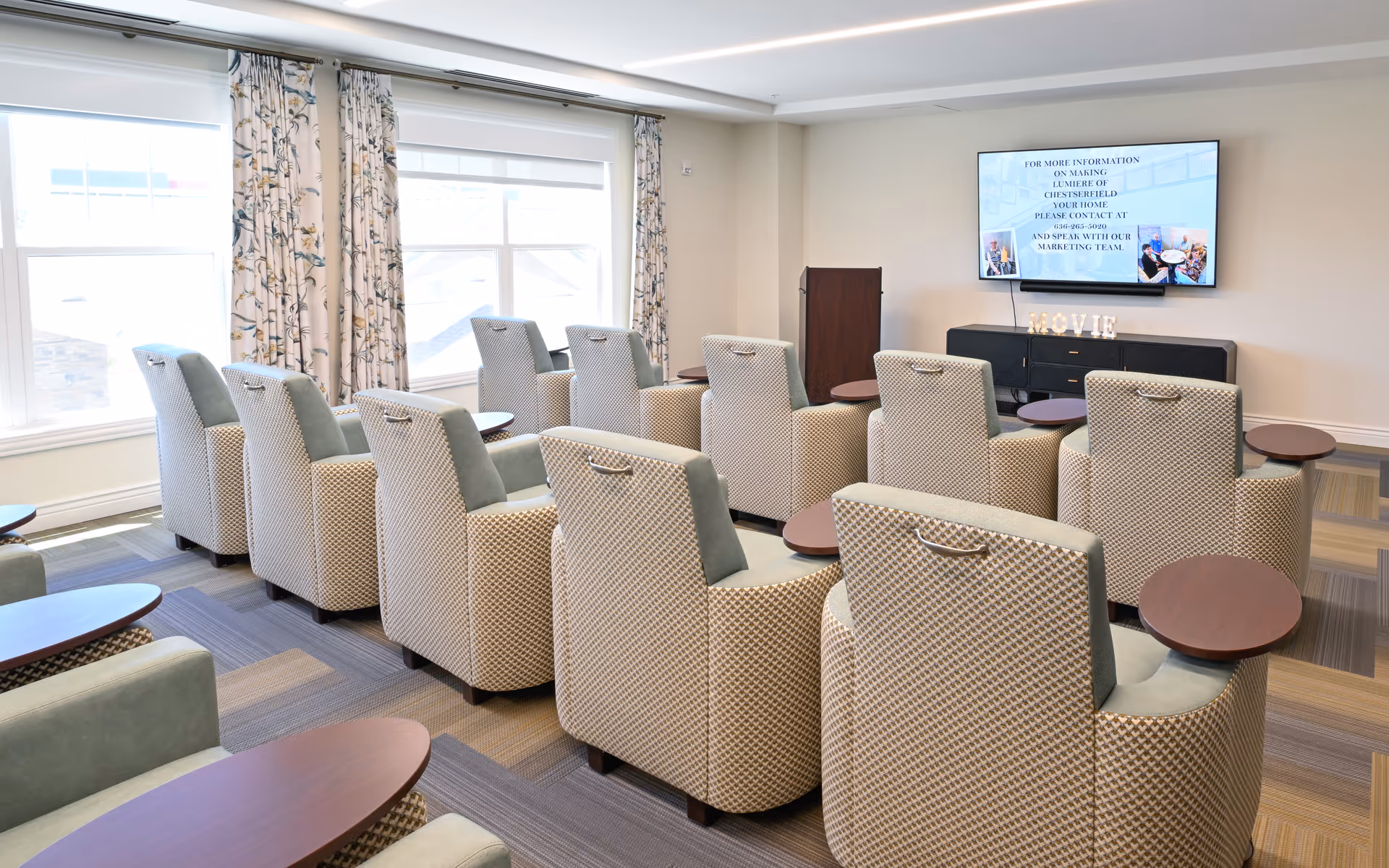 Rows of upholstered armchairs with small round side tables facing a wall-mounted TV in a bright common room.
