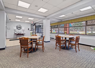 A bright and clean common area with carpeted floor, featuring two wooden tables each surrounded by four wooden chairs. Large windows on the right side let in natural light and offer a view of trees and benches outside. The entrance door and a small reception area with a mirror and decorative items are visible in the background.
