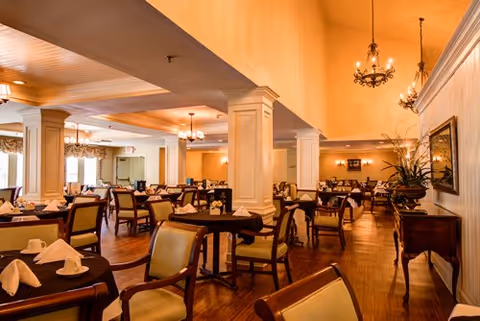 A spacious dining room with multiple tables covered in dark tablecloths, each set with white napkins and cups. The room features wooden floors, cream-colored walls, decorative columns, chandeliers, and large windows allowing natural light to enter.
