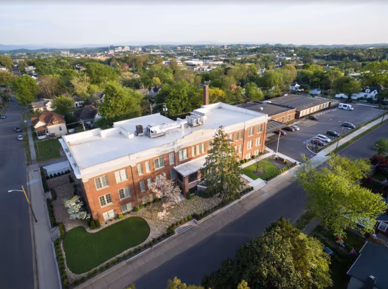 Aerial view of a large brick senior living facility building with a white roof, surrounded by trees and greenery. There is a parking lot adjacent to the building with several parked cars. The facility is located in a suburban area with residential houses and more greenery in the background.