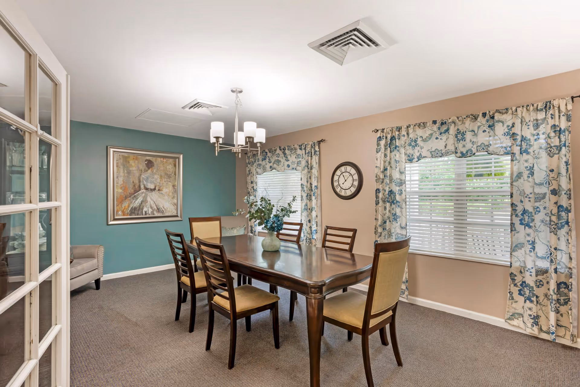 A dining room with a rectangular wooden table surrounded by six chairs with beige cushions. The room has beige walls with one accent wall painted teal, decorated with a framed painting of a woman in a dress. Two windows with floral curtains allow natural light in, and a round wall clock is mounted between the windows. A modern chandelier with five lights hangs above the table, and a vase with flowers is placed in the center of the table.