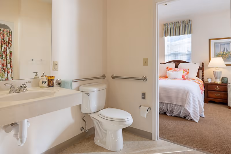 Accessible bathroom with a wall-mounted sink, toilet with grab bars, and an open doorway showing a decorated bedroom.