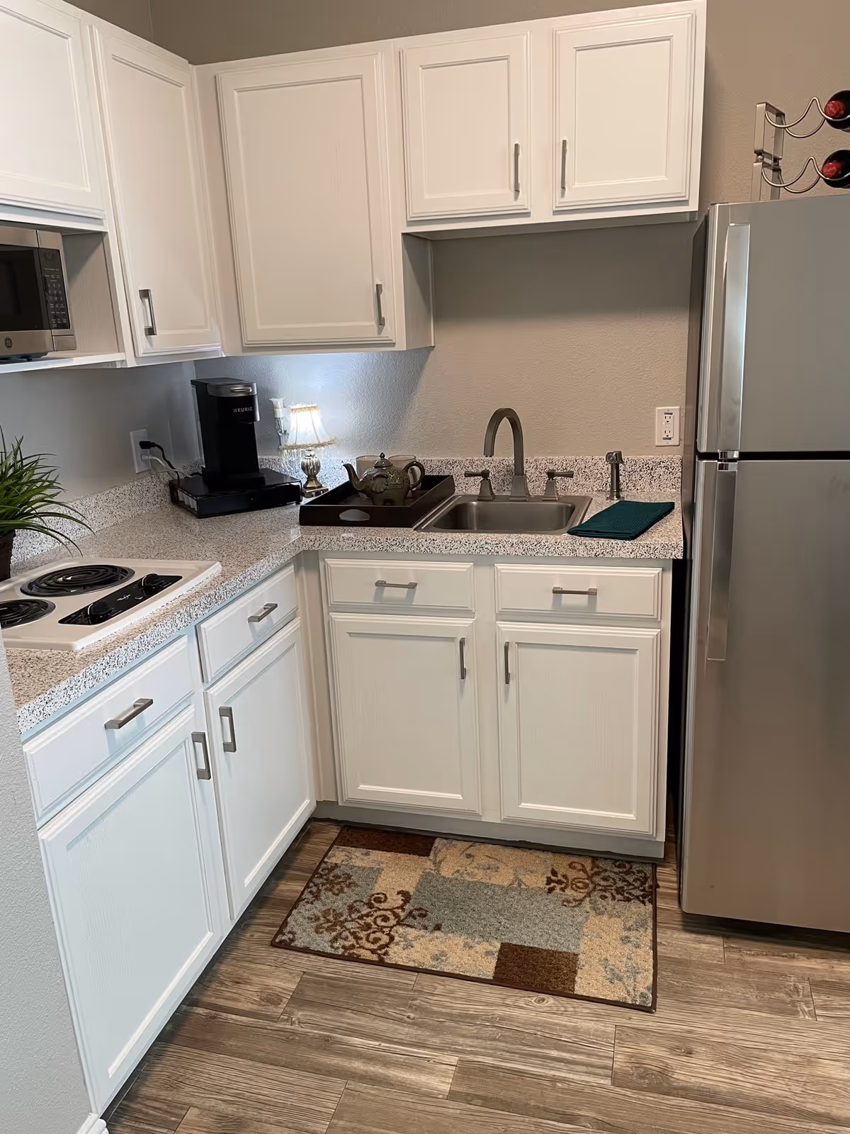 A small kitchen area with white cabinets and drawers, a stainless steel refrigerator, a double sink with a faucet, a coffee maker, a microwave, an electric stovetop with four burners, a decorative tray with a teapot, a small lamp, and a patterned rug on the wooden floor.