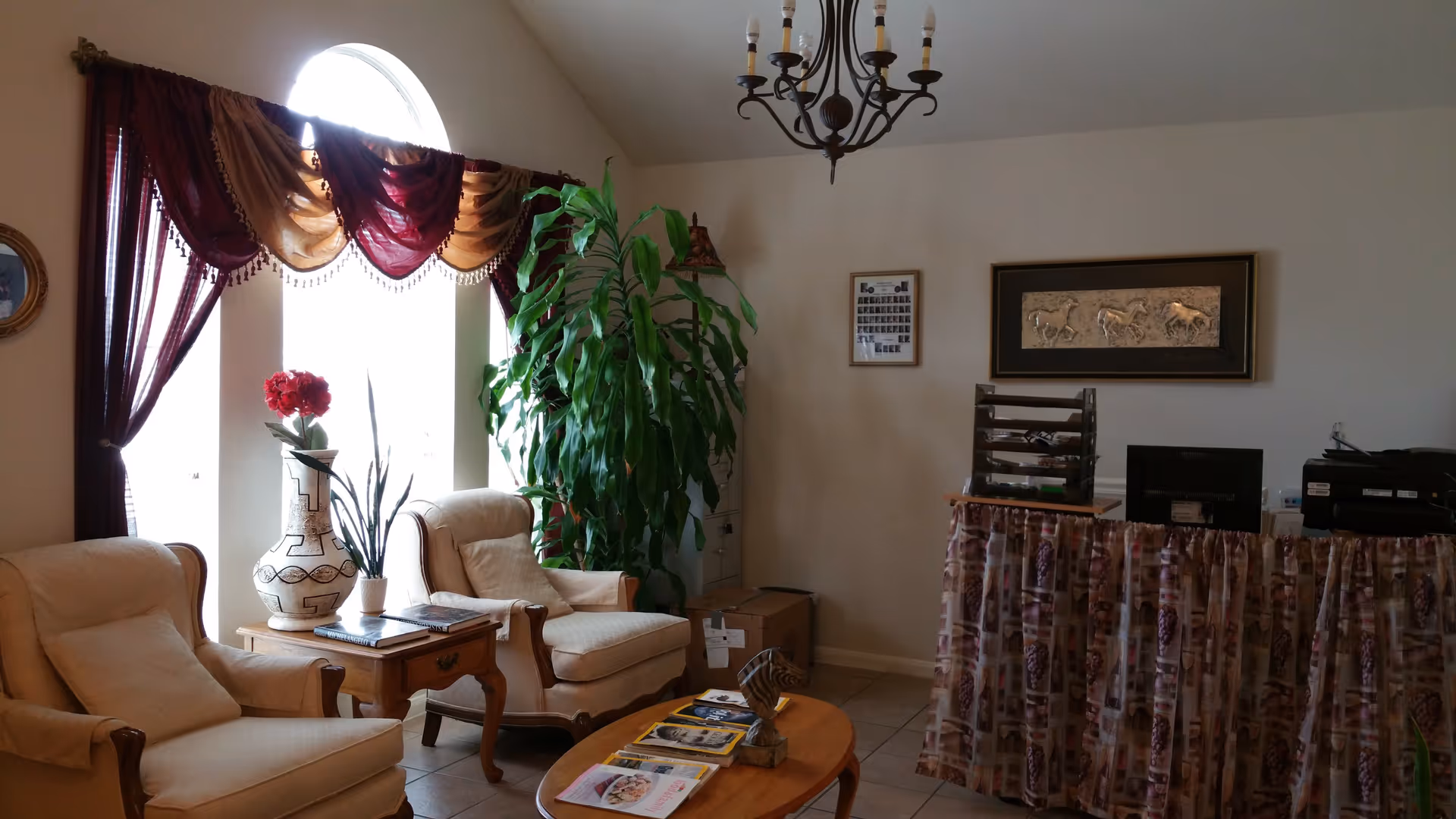 A cozy interior room with two beige armchairs and a wooden side table between them, holding a large decorative vase with red flowers and a smaller plant. A large green plant is in the corner near a window with maroon and gold curtains. A wooden coffee table in front of the chairs has magazines and a small decorative item. On the right side, there is a reception desk with a patterned fabric skirt, a computer monitor, and office supplies. The walls are decorated with framed artwork and a chandelier hangs from the ceiling.
