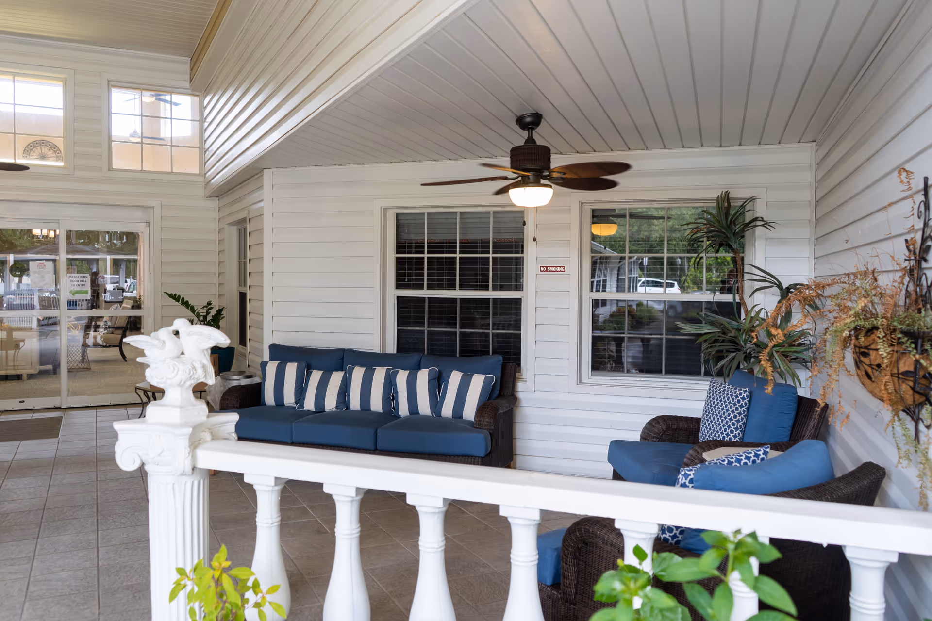 Covered porch area with white railing and tiled floor, featuring a dark wicker sofa and chairs with blue cushions and striped pillows. Ceiling fan with light is mounted on the white paneled ceiling. Windows with white frames and plants decorate the space.