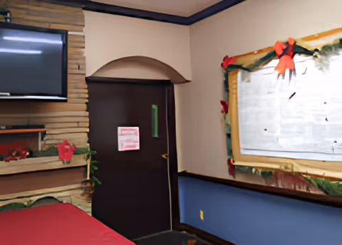 Interior room with a dark brown door, a wall-mounted TV above a stone fireplace, and a bulletin board decorated with holiday garlands and red bows. The walls are painted beige and blue with a dark trim.