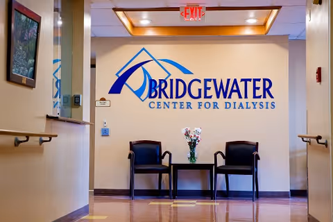 Interior hallway of Bridgewater Center for Dialysis with two black chairs and a small table with a vase of flowers between them. The wall behind has the Bridgewater Center for Dialysis logo. Handrails are mounted on both sides of the hallway, and an exit sign is visible on the ceiling.