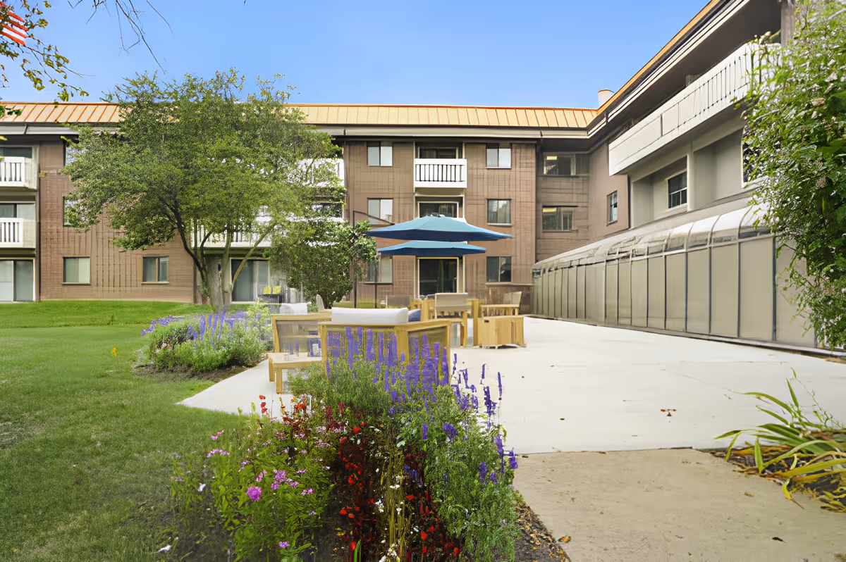 Courtyard patio with seating and a blue umbrella, flower beds, and the multi-story senior living building in the background.
