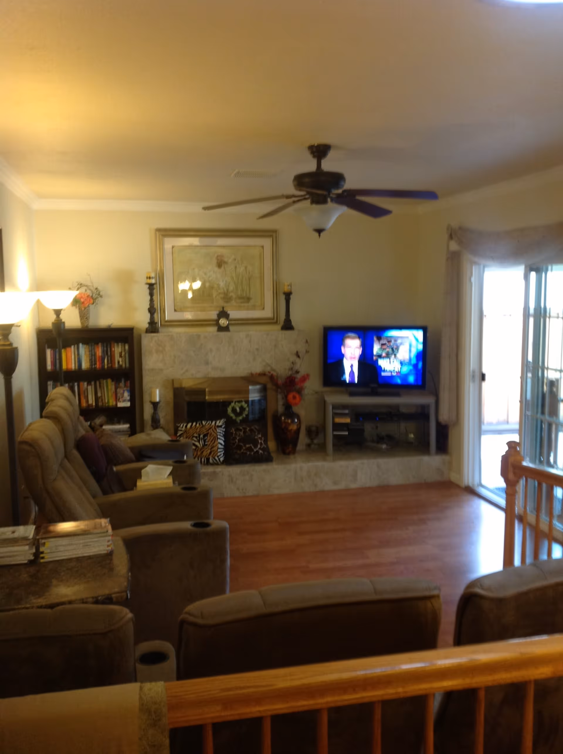 A cozy living room with beige walls and wooden flooring. There are several cushioned recliner chairs arranged facing a TV on a stand. The TV is on, showing a news anchor. Behind the chairs is a wooden railing. On the far wall, there is a fireplace with decorative pillows on the hearth, a large framed floral painting above it, and two tall candle holders on either side. To the left of the fireplace is a bookshelf filled with books and a lamp with a flower vase on top. To the right, there is a sliding glass door with curtains, letting in natural light.