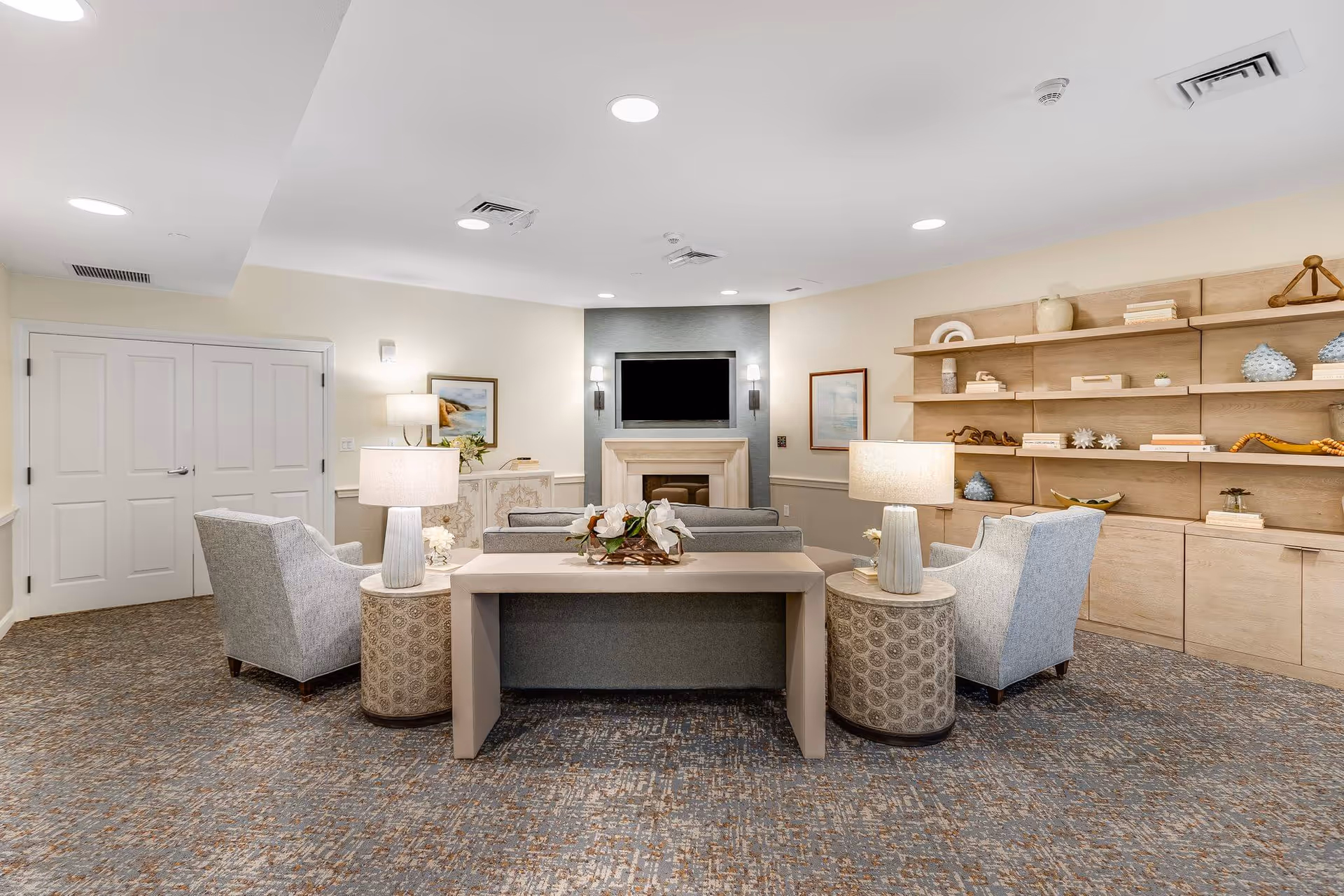 A cozy living room area in a senior living facility with a gray sofa and two gray armchairs arranged around a fireplace with a mounted flat-screen TV above it. There are two round side tables with lamps on either side of the sofa, a rectangular console table behind the sofa with a floral arrangement, and a large wooden shelving unit on the right wall decorated with books and ornaments. The room has recessed ceiling lights and framed artwork on the walls.