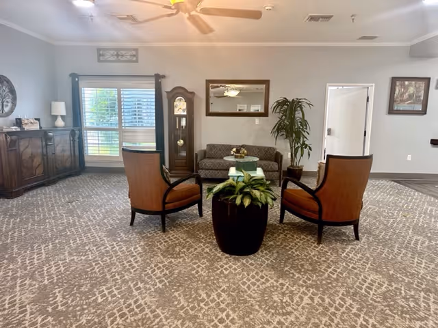 A spacious living room area with patterned carpet, two orange cushioned chairs facing a small glass coffee table with a plant on it, a gray sofa against the wall, a tall grandfather clock, a large potted plant, a window with blinds, a ceiling fan, and framed artwork on the walls.