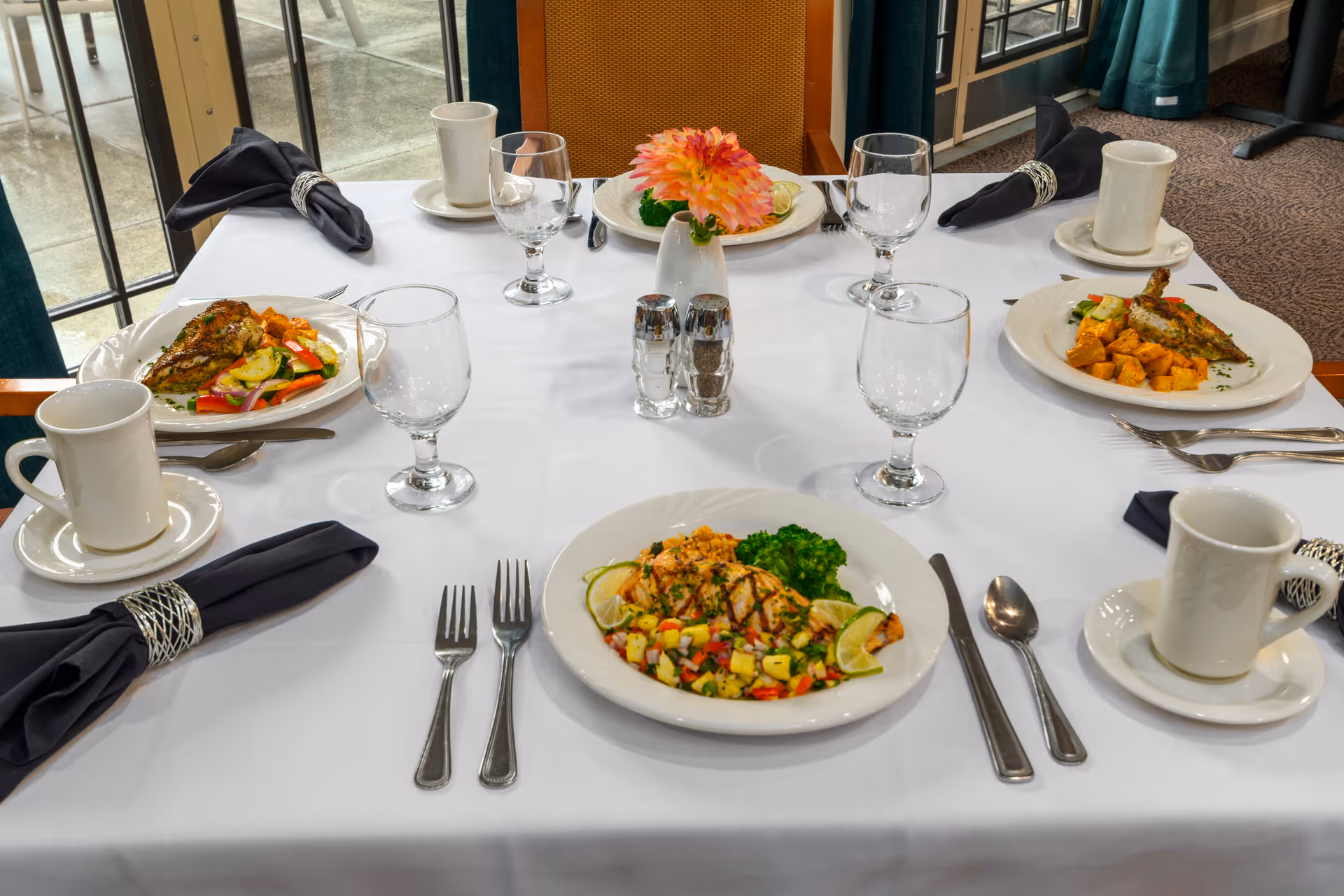 A dining table set for four with white tablecloth, black napkins with silver napkin rings, white plates with meals including grilled chicken, vegetables, and broccoli, clear water glasses, white coffee cups on saucers, silverware, a small vase with a pink flower, and salt and pepper shakers. The table is near large windows with a view of an outdoor patio.