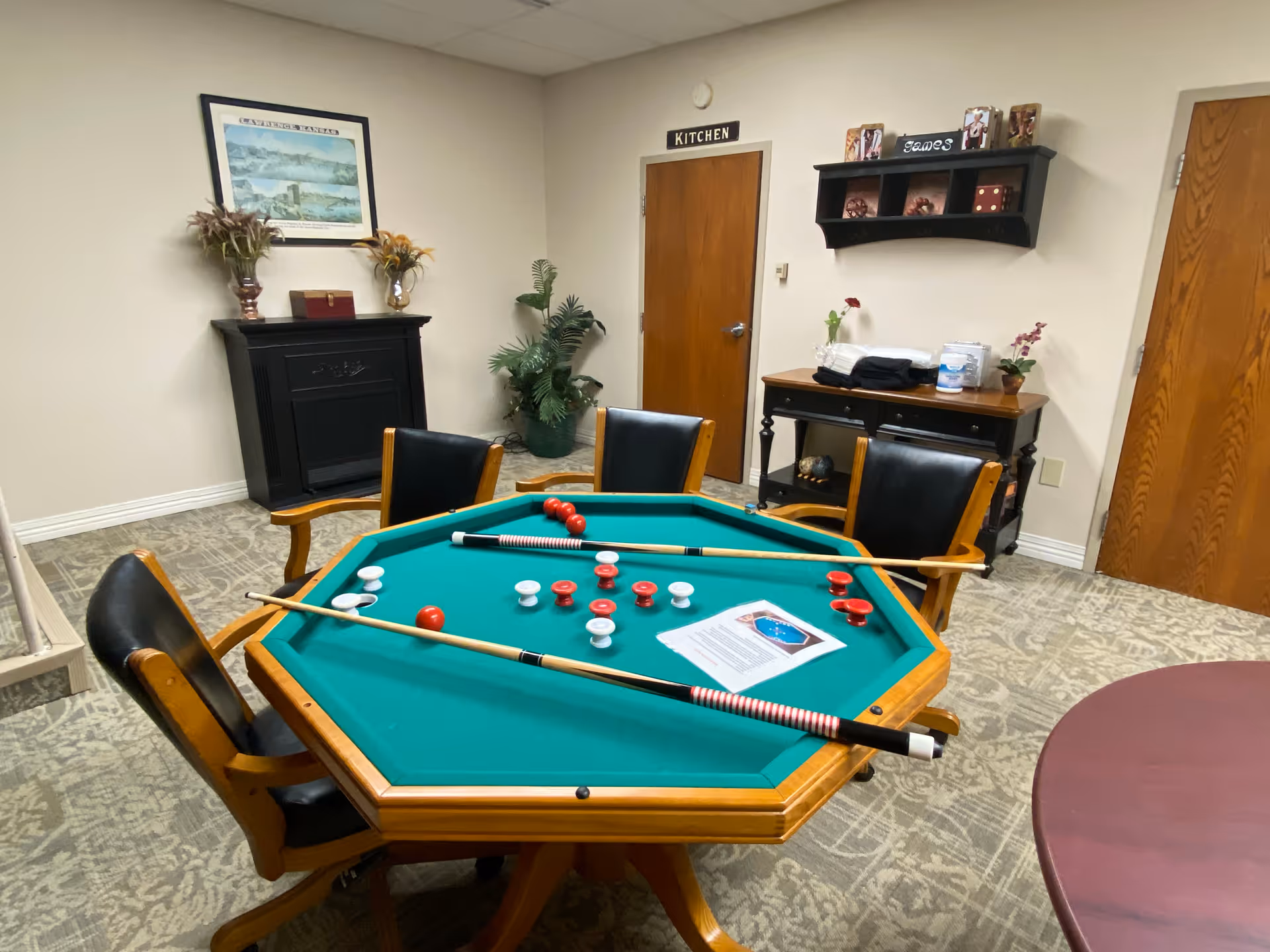 A game room with an octagonal green felt game table surrounded by four wooden chairs with black cushions. The table has red and white game pieces and two cue sticks on it. In the background, there is a door labeled 'KITCHEN', a black fireplace mantel with decorative items, a small table with plants and supplies, and a wall shelf with decorative dice and a sign that says 'Games'.
