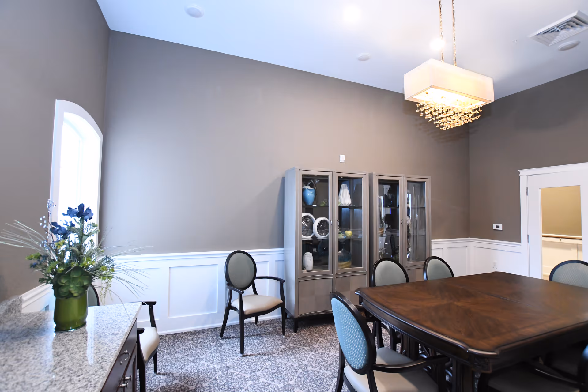 Dining room with a large wooden table surrounded by upholstered chairs, glass-front cabinets, a floral arrangement, and a chandelier.