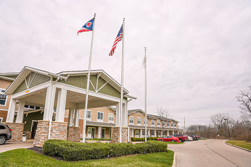 Exterior view of Traditions of Beavercreek senior living facility showing the front entrance with a covered drop-off area, three flagpoles with flags including the Ohio state flag and the American flag, a row of parked cars, and a cloudy sky.