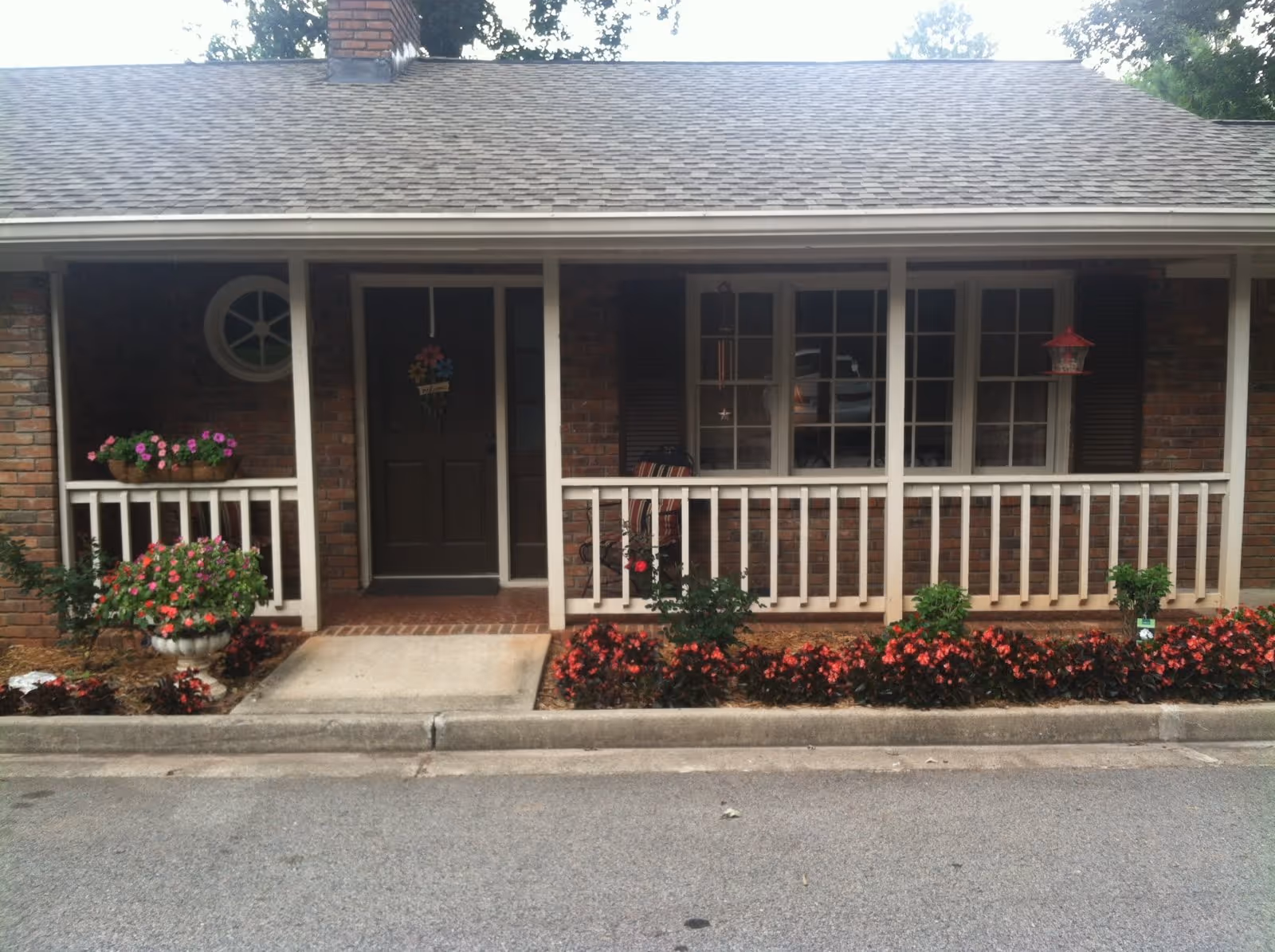 Brick single-story building front with a covered porch, white railing, central front door, windows, and flower beds along the curb.