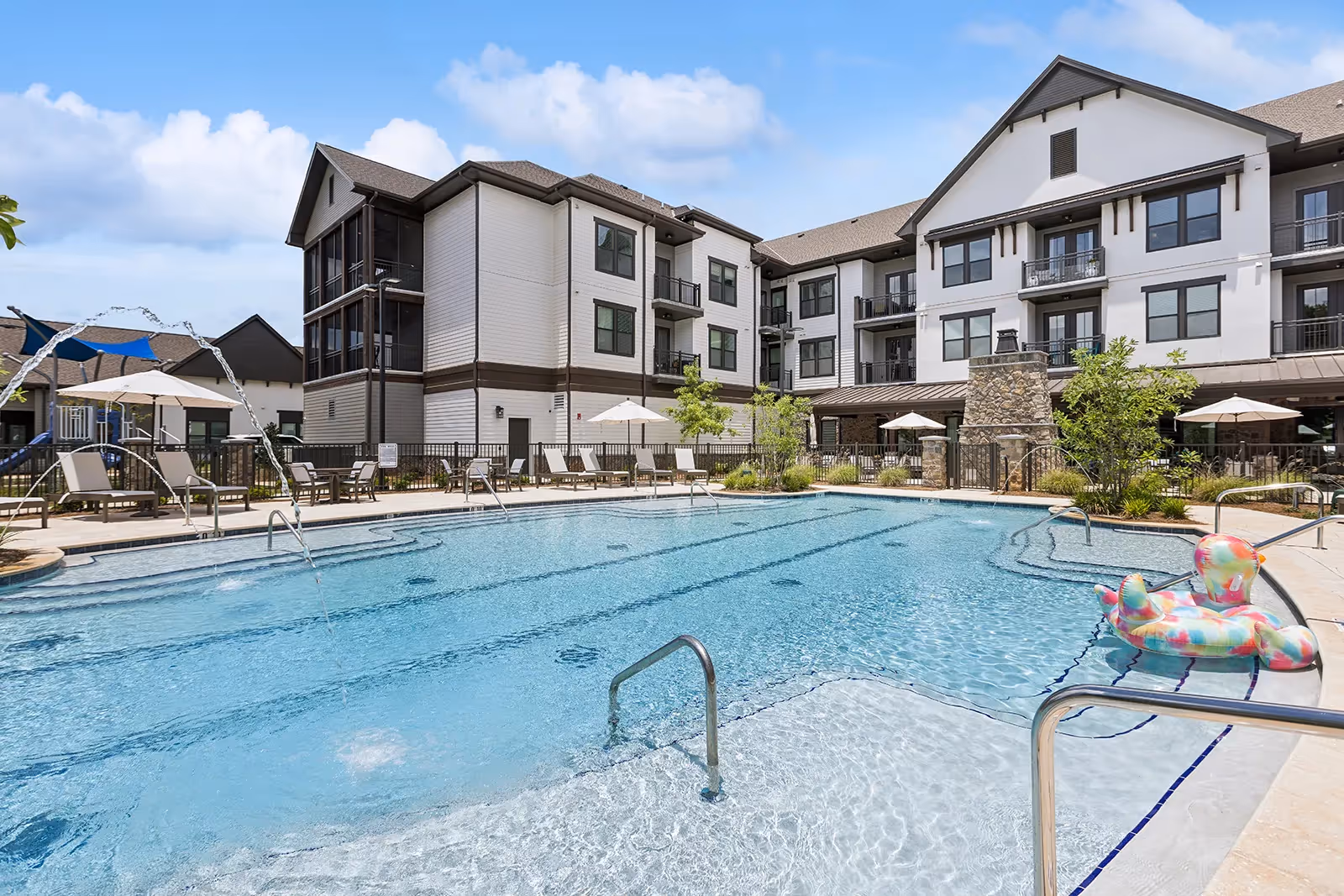 Outdoor swimming pool with clear blue water in front of a multi-story senior living facility building. The pool area features lounge chairs, umbrellas, water fountains, and a colorful inflatable duck float. The building has white siding with dark trim and balconies overlooking the pool.