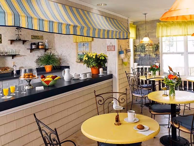 A cozy dining area with yellow round tables and black wrought iron chairs. The tables have small flower vases and some have coffee cups and pastries. There is a counter with a striped yellow and blue awning above it, displaying glasses of orange juice, a bowl of fruit, a cake stand with pastries, and a potted plant. The room has large windows letting in natural light and yellow curtains.