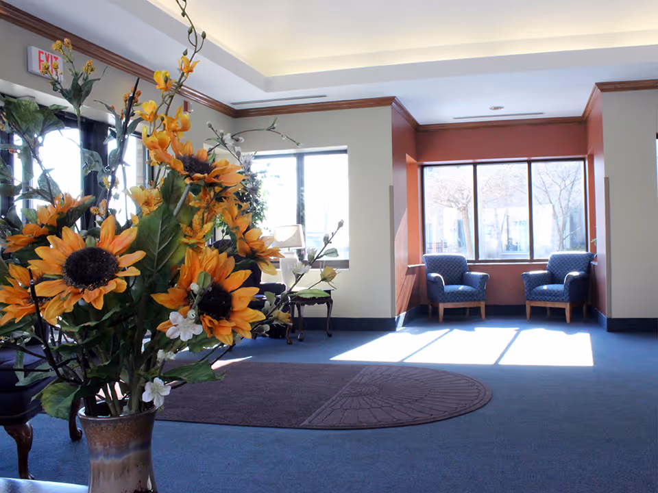 A bright and spacious common area with large windows letting in natural light. The room features two blue upholstered armchairs near the windows, a round brown rug on blue carpeted floor, and a vase with vibrant yellow sunflowers and other flowers in the foreground. The walls are painted in neutral tones with a reddish accent wall around the window area.