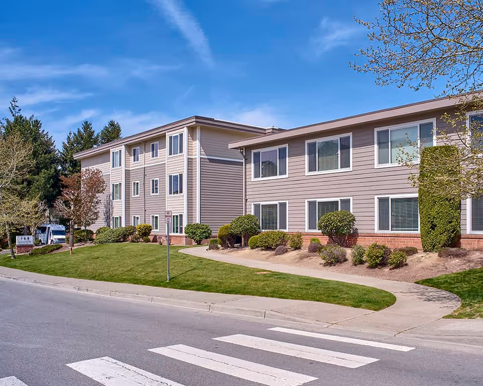 Front exterior view of a two- and three-story senior living building with landscaped lawns, a sidewalk, and a crosswalk under a blue sky.