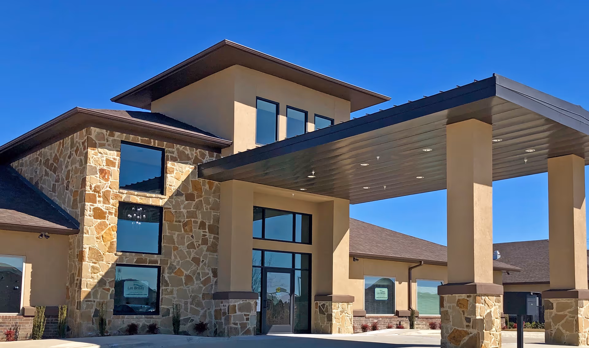 Exterior view of Las Brisas Rehabilitation and Wellness Suites building featuring a stone and stucco facade, large windows, and a covered entrance with pillars under a clear blue sky.