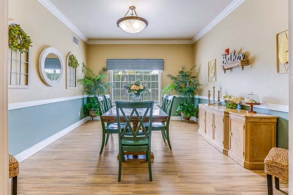 A bright dining room with a wooden table and six green chairs. The room features light wood flooring, beige and green walls with white trim, and a large window with blinds and a blue valance. There are decorative plants in the corners, wall mirrors, and framed leaf artwork. A wooden sideboard holds candles, flowers, and a cake stand, with a wall decoration that says 'Gather' above it. A ceiling light fixture hangs above the table.