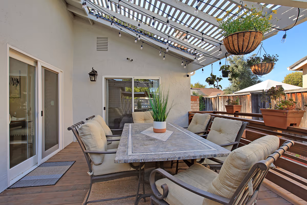 Outdoor patio area with a rectangular stone table surrounded by cushioned chairs under a pergola with hanging string lights and several hanging and potted plants. Sliding glass doors lead inside the building.