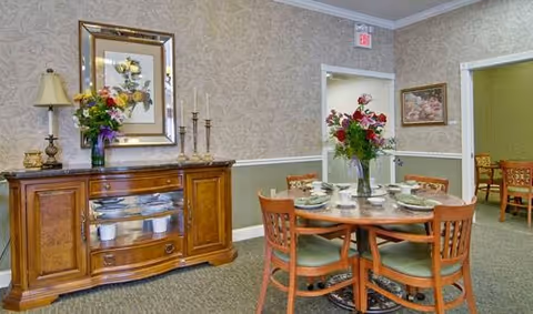 A dining area in a senior living facility featuring a round wooden table set with plates, bowls, and utensils, surrounded by four wooden chairs with green cushions. A vase with red and white flowers is placed in the center of the table. To the left, there is a wooden sideboard with a lamp, a floral arrangement, and decorative candlesticks. The walls are decorated with floral wallpaper and framed artwork. Another dining table and chairs are visible in an adjacent room through an open doorway.