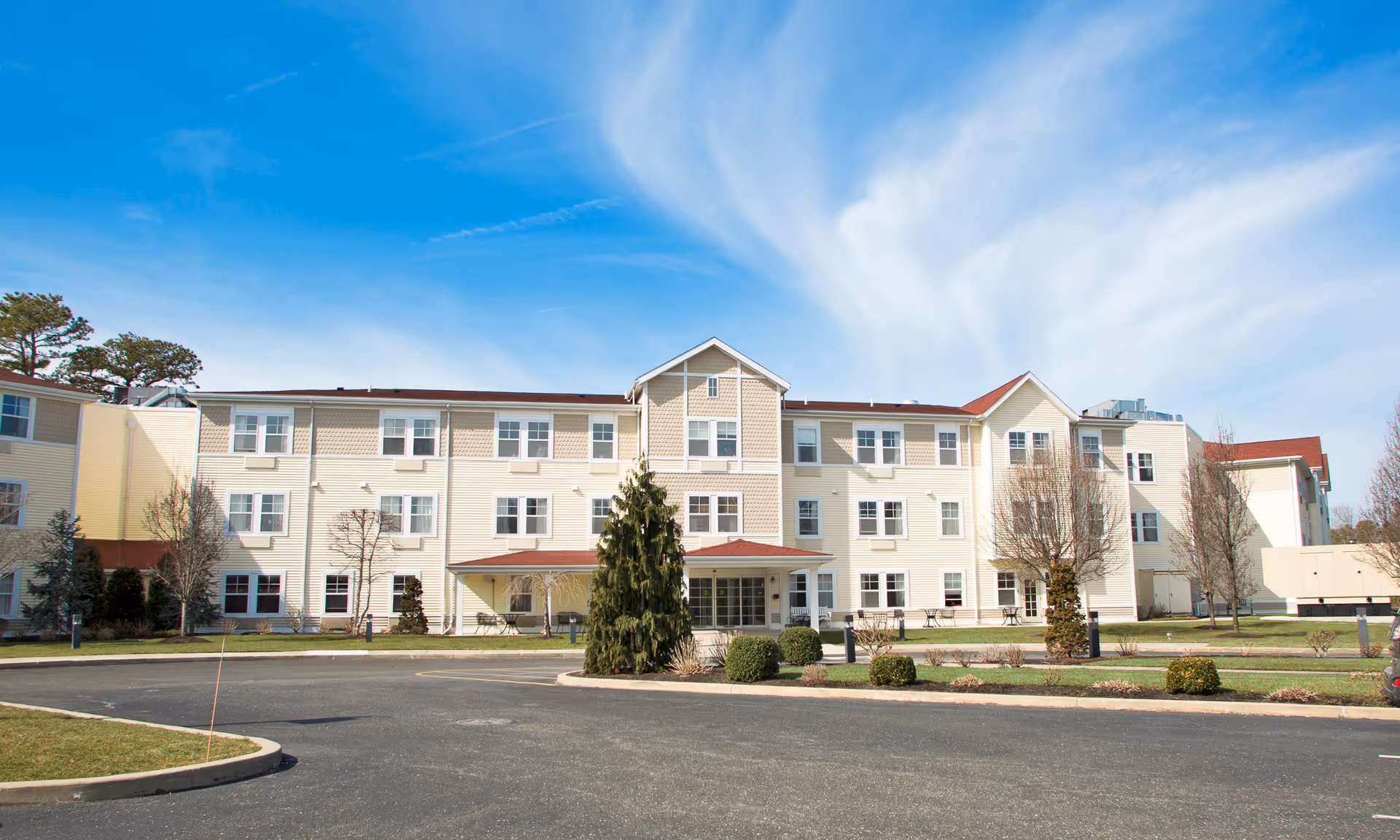 Three-story beige senior living building with a central entrance, landscaped grounds, and a bright blue sky.