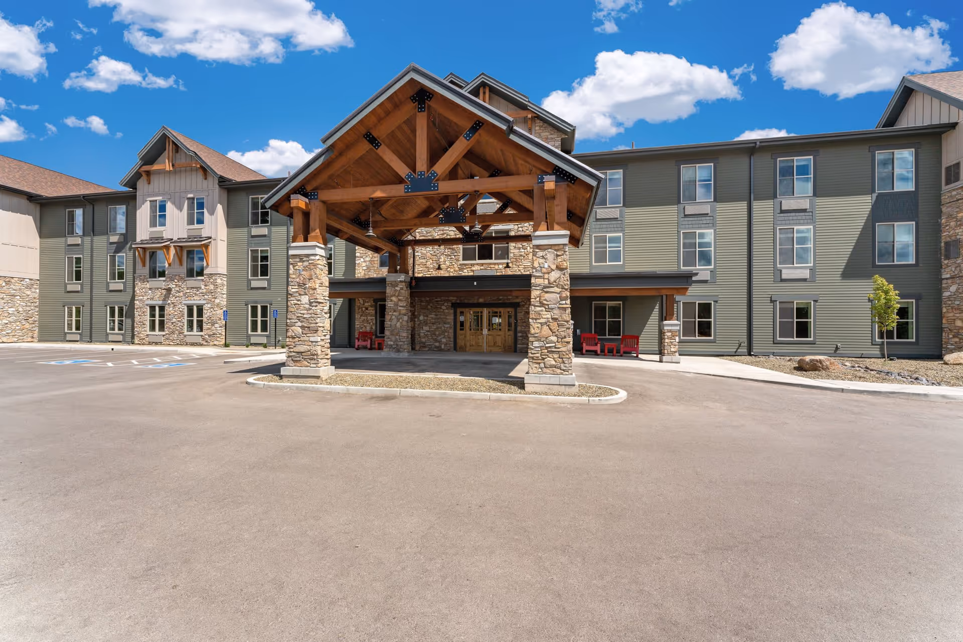 Front exterior view of The Bluffs of Flagstaff Senior Living building with a large covered entrance supported by stone pillars, multiple windows, and a clear blue sky with scattered clouds.