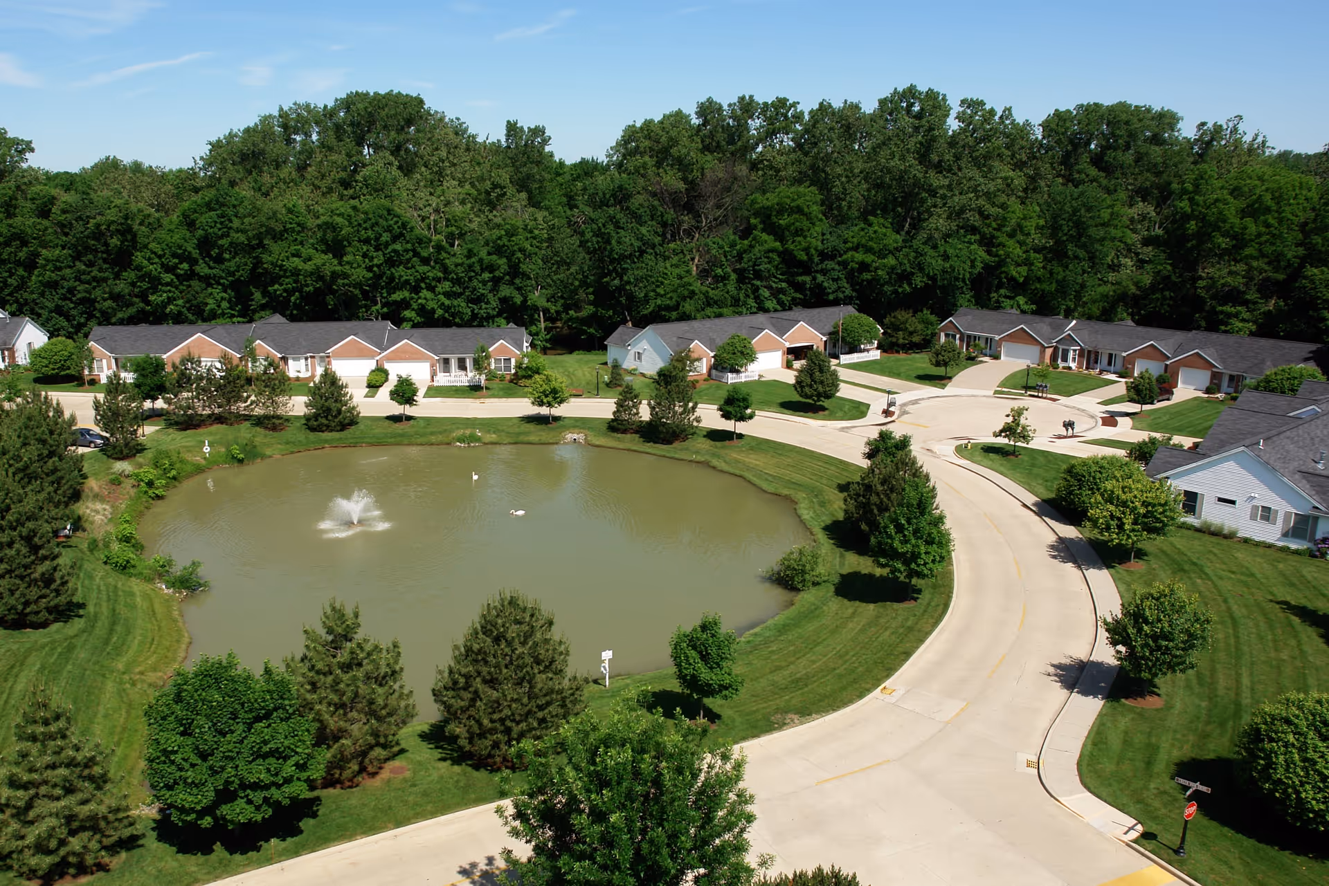 Aerial view of a senior living community at Ohio Living Swan Creek featuring a circular pond with a fountain in the center, surrounded by well-maintained lawns, trees, and single-story residential buildings with garages. The area is bordered by a dense forest in the background under a clear blue sky.