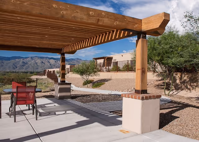 Outdoor patio area with a wooden pergola casting shadows on the concrete floor, a table with red chairs, desert landscaping with gravel and sparse trees, and mountains in the background under a partly cloudy sky.