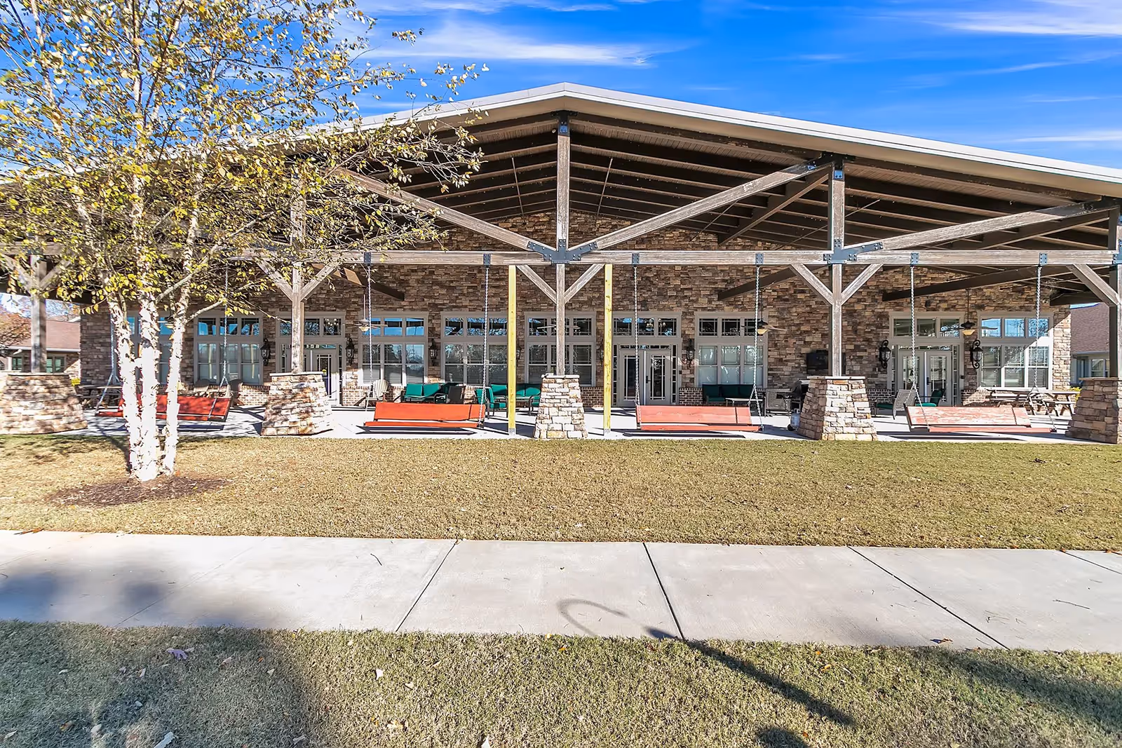 Outdoor covered patio area with wooden beams and stone pillars, featuring several red wooden swings and green cushioned seating. The patio is attached to a building with large windows and stone exterior walls. There is a tree on the left side and a sidewalk in front of the grassy area.