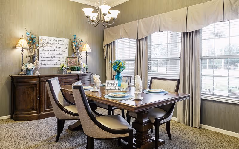 A well-lit dining room with a wooden dining table set for four, featuring plates, cups, and napkins. The room has large windows with beige curtains allowing natural light to enter. A wooden sideboard against the wall is decorated with lamps, flowers, and framed artwork. The walls are covered with textured wallpaper, and a chandelier hangs above the table.