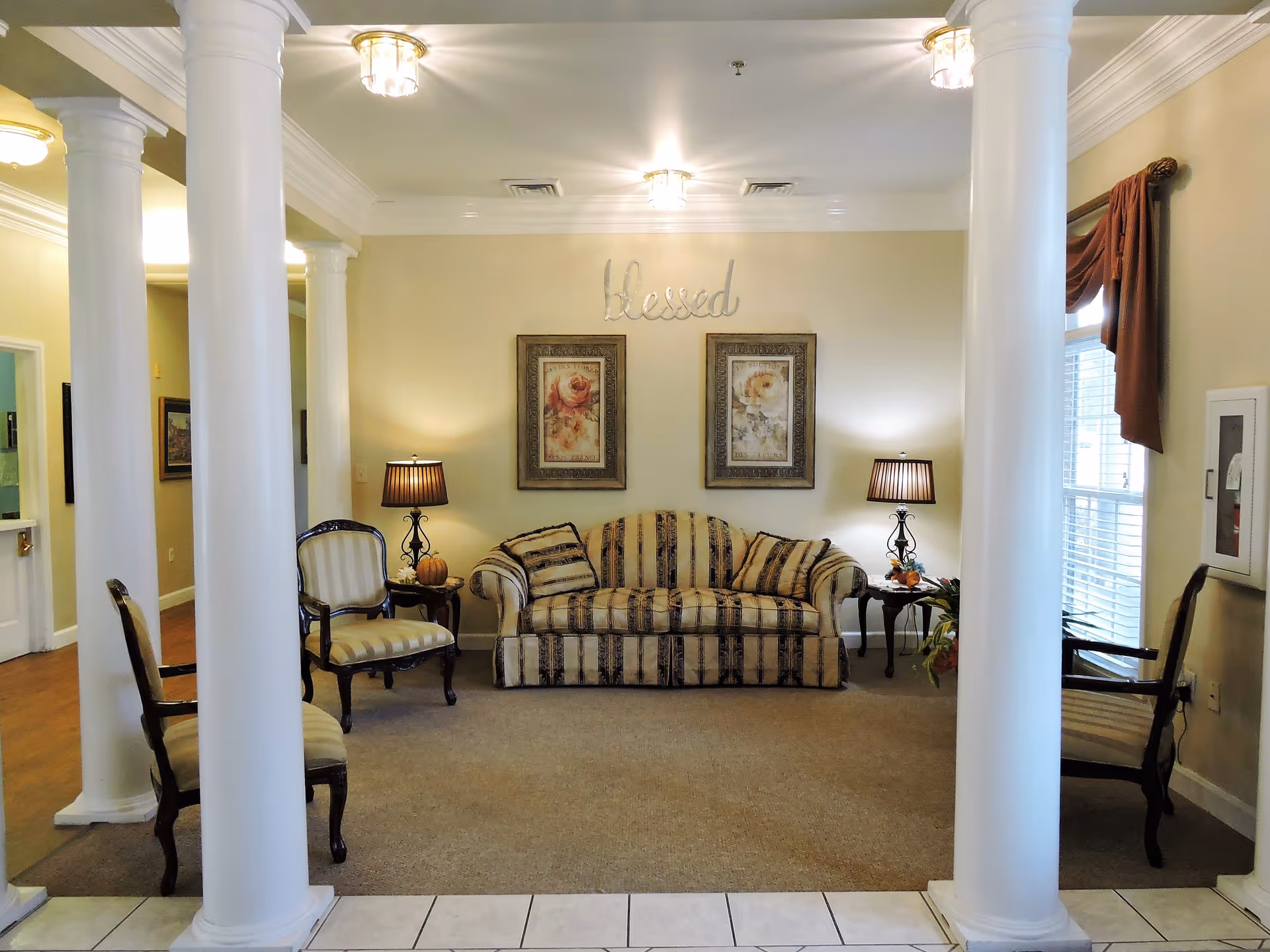 Cozy common room with a striped sofa, armchairs, side tables and lamps framed by white columns and floral artwork under a 'Blessed' wall sign.