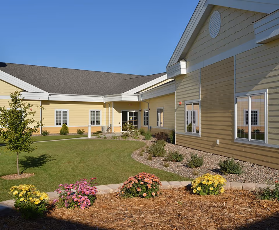 Yellow single-story senior living building with a landscaped lawn, flowerbeds, and a clear blue sky.
