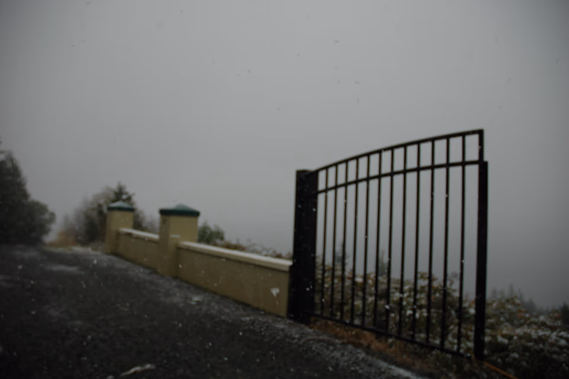 A black metal gate opens beside a low stucco wall along a snowy, foggy driveway.